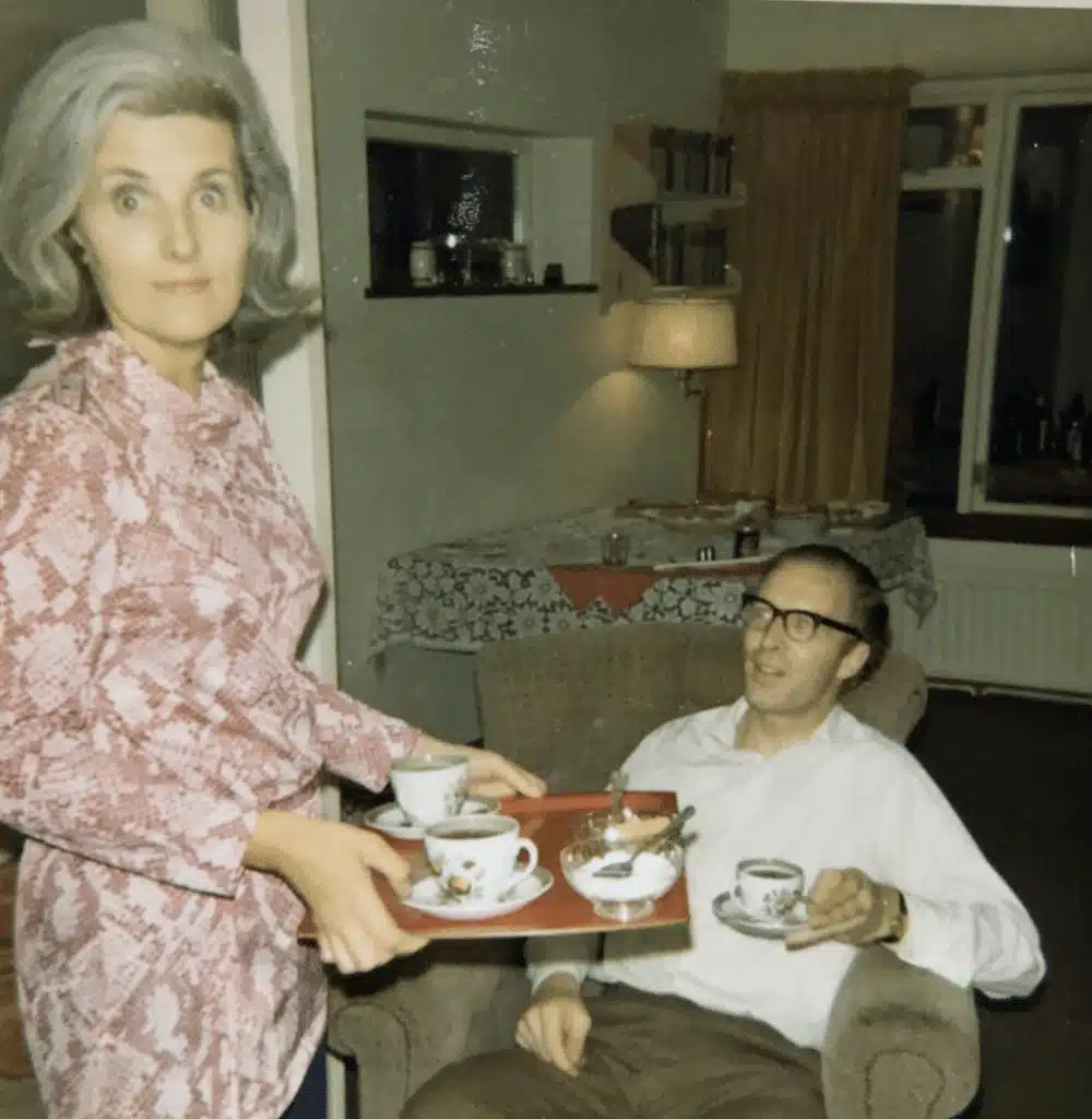 Vintage photo of a Ave serving tea on a tray to a seated man in a living room, with a patterned tablecloth and bookshelves in the background