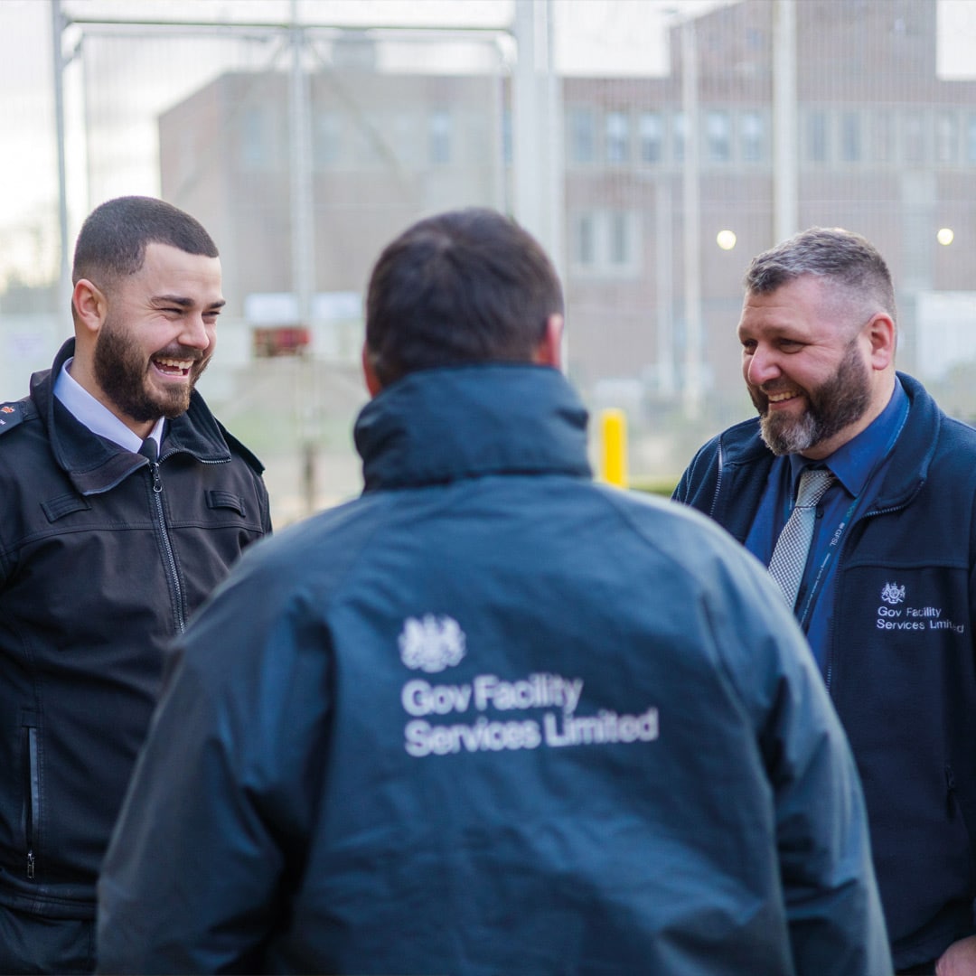 Alt text:
Three Gov Facility Services Limited employees wearing branded uniforms talk and laugh together in a secure outdoor setting, reflecting a positive and unified team culture as part of an internal communications and employer brand project