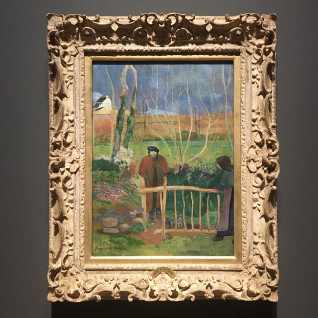 Framed painting by Gauguin of two figures standing near a rustic wooden gate in a lush green landscape, with bare trees and a house in the background.
