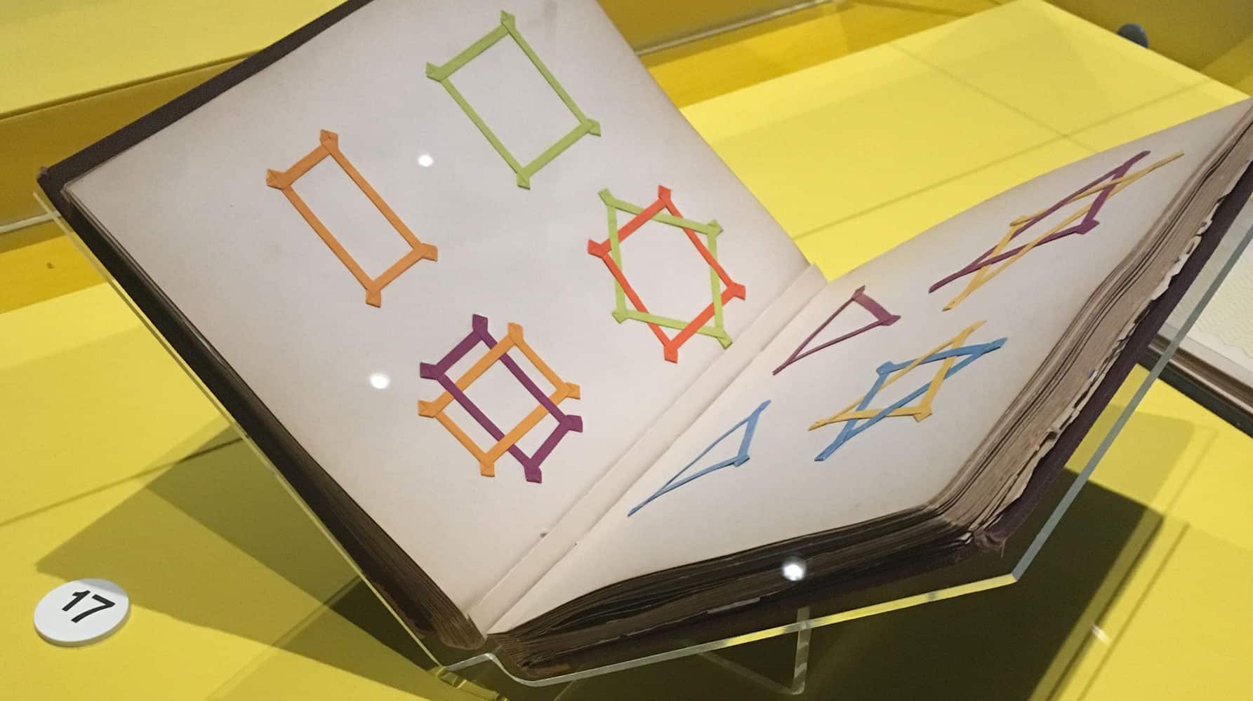 Open book on display showing colourful geometric shapes made of ribbon arranged in square and star-like patterns.