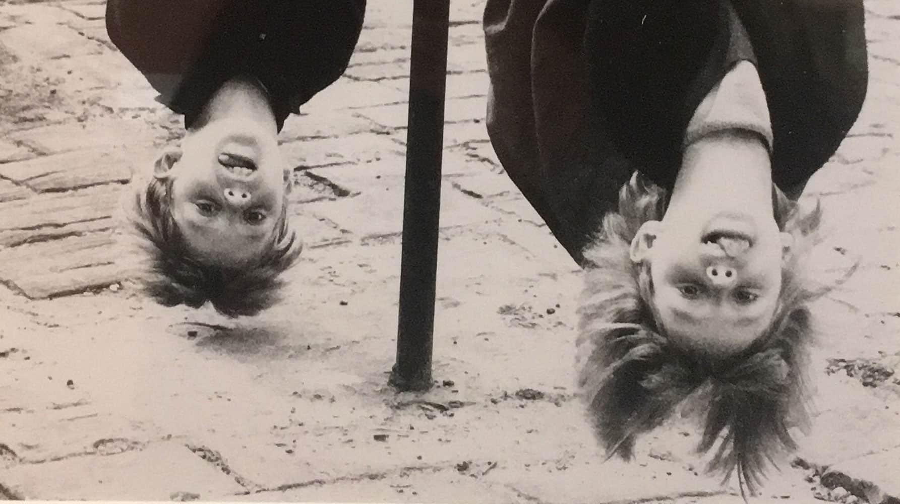 Black and white photo of two children hanging upside down on a pole, their hair dangling and tongues out playfully.