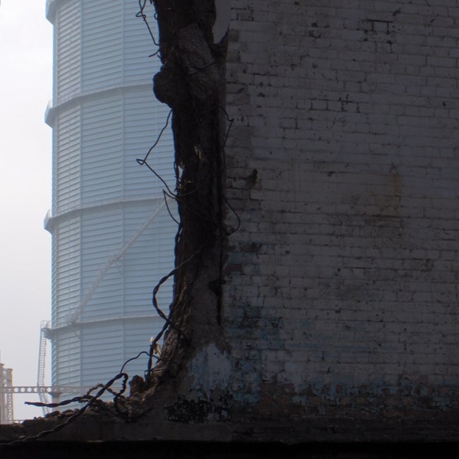 View through a broken brick wall framing a distant gas holder.