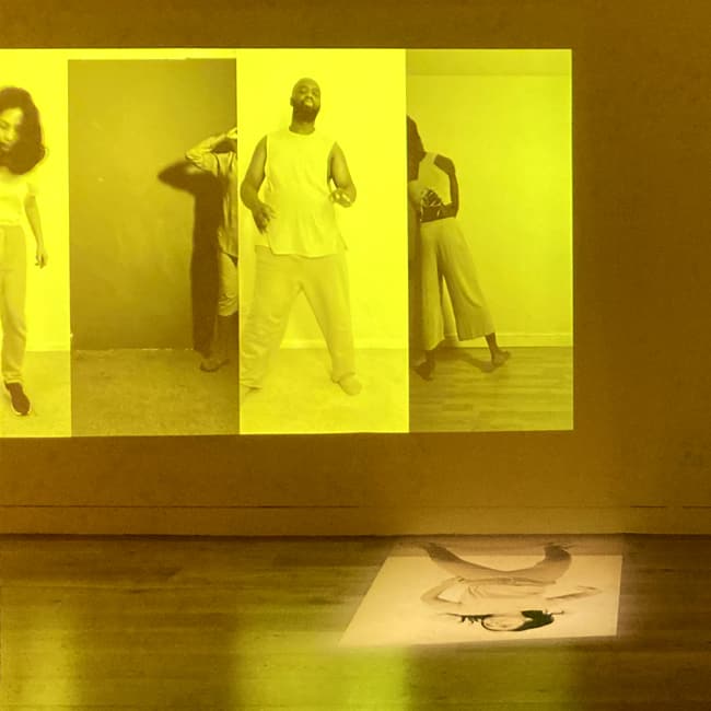 Video projection of four dancers performing individually on a yellow background, with an additional projection of a dancer on the floor below.