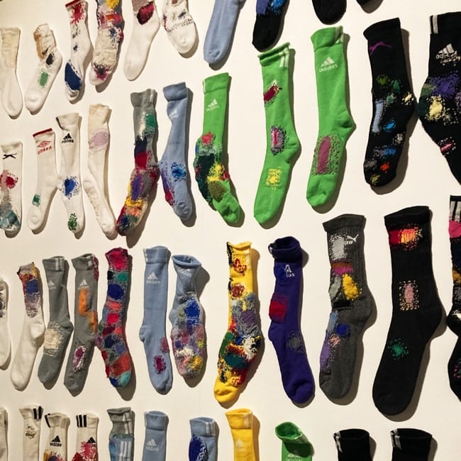 Wall-mounted art installation of colourful socks from various brands, visibly repaired with multicoloured stitched patches.