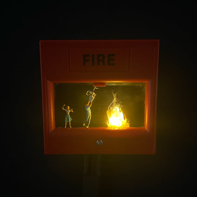 Miniature scene of people trying to put out a fire inside a fire alarm box.