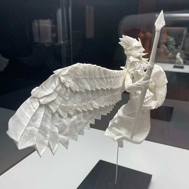 Intricate white origami sculpture of a winged figure holding a spear.