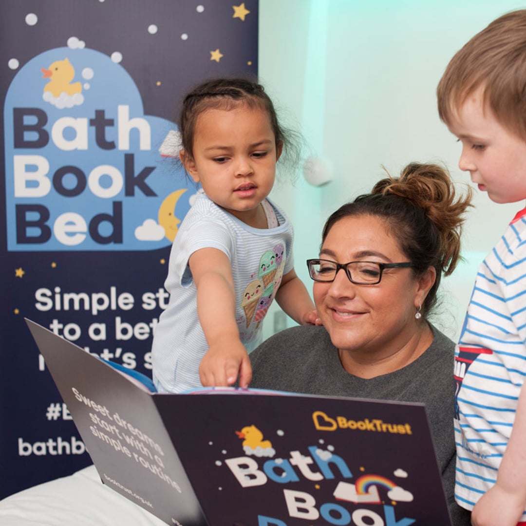 A parent sits with two children on a bed reading a large “Bath Book Bed” booklet, with a campaign banner in the background promoting BookTrust’s simple bedtime routine.